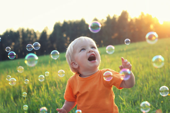 Cute toddler blond boy playing with soap bubbles on summer field. Happy childhood concept. Authentic lifestyle image