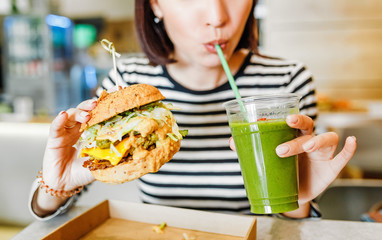 A young woman drinks green smoothies and eats a burger in a vegan fast food restaurant - obrazy, fototapety, plakaty