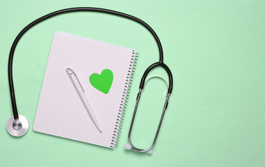 Notepad with pen, decorative heart, stethoscope on pink pastel background, medical concept, top...