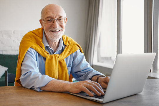 Picture of successful positive elderly bearded European travel blogger typing article on portable computer, looking and smiling at camera, wearing stylish sweater around neck over blue shirt