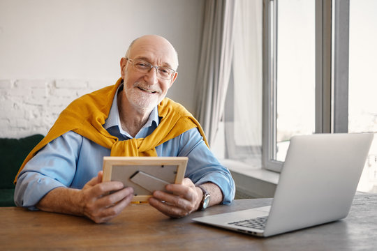 Portrait of positive successful attractive senior man employee with gray beard working in modern office interior, using laptop, holding photo frame and smiling while missing his grandchildren