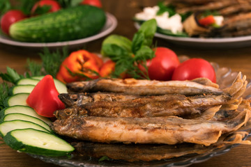 Small fish (capelin), completely fried to a ruddy crisp, laid on a glass plate with sliced cucumbers, sweet pepper and tomatoes.