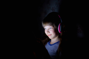 the child in wireless headphones listens to music at night under a blanket and looks at the tablet - Powered by Adobe