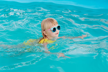 Little girl in a yellow swimsuit and sunglasses is swimming and jumping to swimming pool at resort. Summer fun in the pool. 