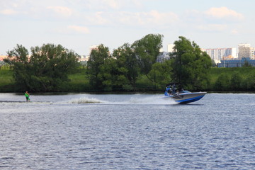 Obraz premium Gray-blue towing boat rides wakeboarder on the river in summer on the background of water and shore with green trees