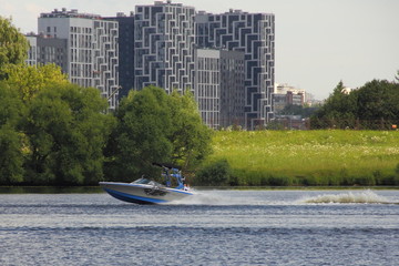 Obraz premium Silver-blue towing boat fast floating on the river in summer on the background of water, shore with green trees and modern city building in Moscow, Strogino