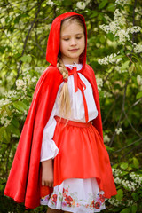 Smiling blond girl posing in a dress of little red riding hood near green trees