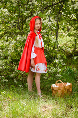 Smiling blond girl posing in a dress of little red riding hood near green trees