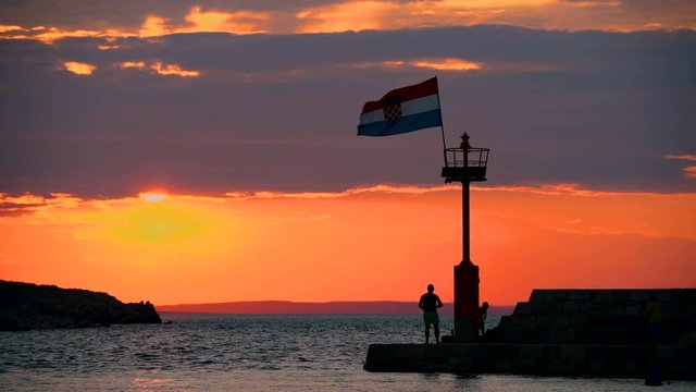 Stone pier with lighthouse and flying Croatian flag in sunset, people walking and sitting on stone wall, silhouettes of people only, individuals not recognizable