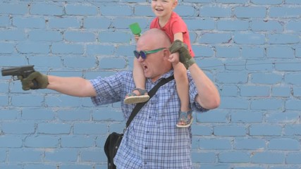 Elderly man with shaved head wearing sunglasses is holding small child and gun in his hands. boy is holding child's shovel. man shoots around at enemies. Taking care of your child's safety