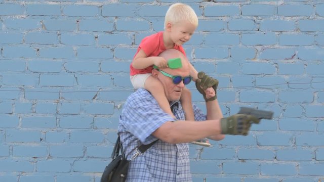 Elderly man with shaved head wearing sunglasses is holding small child and gun in his hands. boy is holding child's shovel. man shoots around at enemies. Taking care of your child's safety