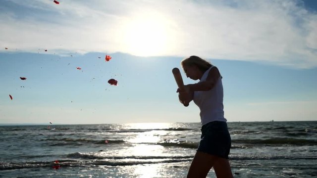 slow motion: young sexy girl beats a baseball bat with vegetables and a tomato fruit. It is located on the beach, sandy beach
