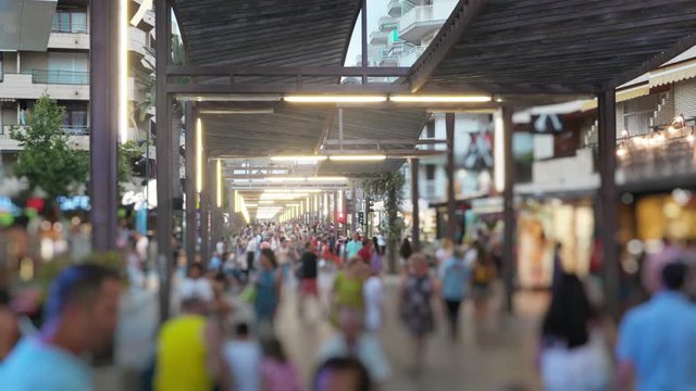 Slow motion of busy shopping street on summer resort in the evening. Unfocused crowd of tourists strolling along shimmering shopwindows in Salou, Spain.