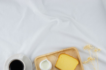 A cup of coffee, milk and butter cake on wooden plate with flower on white bed with copy space