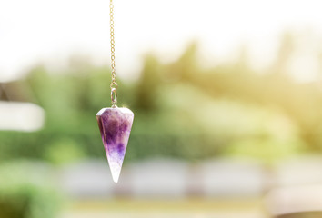 Purple Amethyst crystal pendulum on chain, outdoors trees and nature on the background. - obrazy, fototapety, plakaty
