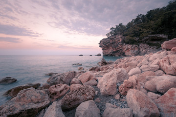 Beautiful rocky beach at sunset time. Big textured boulders on foreground and cliffs, covered with pine trees on background. Antalya. Turkey.