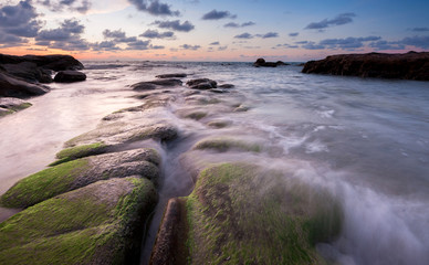 Obraz premium view of beautiful sunset at the beach with natural coastal rocks covered by green moss. soft focus due to slow shutter effect.