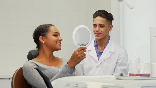 Young happy beautiful African woman smiling, checking her perfect healthy teeth in the mirror, after dental examination at the dentist office. Professional dentist working with his patient.