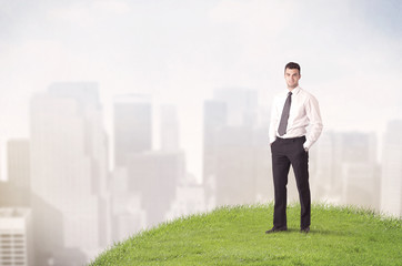 A cheerful caucasian elegant sales manager standing in small green grass in front of faded city landscape, tall buildings concept.