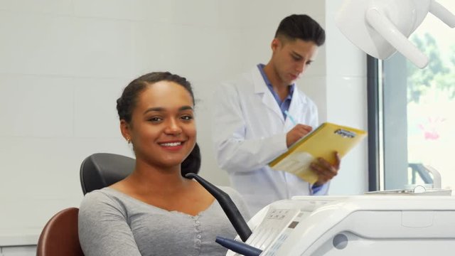 Young beautiful happy African woman examining her perfect healthy smile in the mirror, her dentist filling medical papers on the background. Medicine, healthcare, teeth concept.