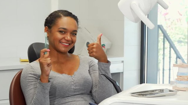 Gorgeous happy African woman smiling with her perfect white teeth, showing thumbs up. Beautiful young woman with healthy flawless smile holding a toothbrush, posing a the dental clinic.