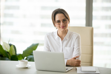 Smiling confident millennial businesswoman posing at personal desk, happy successful female ceo smiling at camera, photo portrait of business owner in small modern office workplace in business center