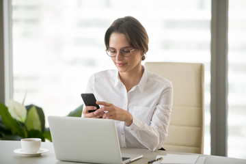 Happy successful businesswoman scrolling news feed in app or checking social media on smartphone, confident female ceo reading news or texting on cell, having coffee at desk in business office center
