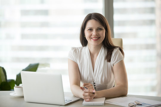 Portrait of smiling middle aged businesswoman working at laptop at office desk, making notes at workplace, confident successful female worker looking at camera, posing for company business photo