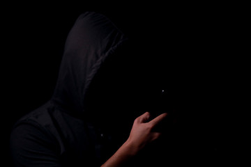 Silhouette of the unknown person in a hood with the mobile phone in a hand in the dark. Cyber...