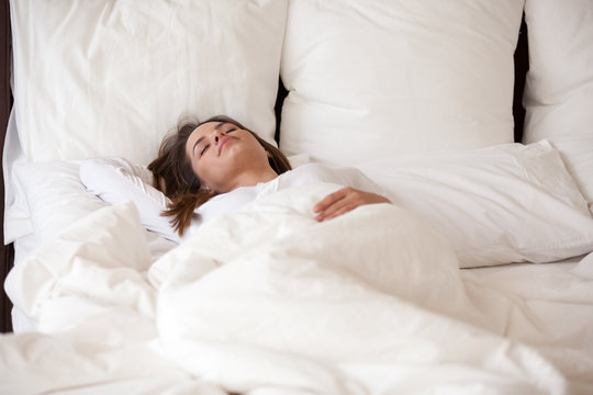 Young female sleeping well on soft pillows covered with warm duvet, asleep woman resting in white cozy bedroom on good quality mattress, taking nap, calm girl relaxing in big comfy bed, seeing dreams