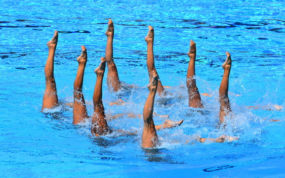 Synchronized Swimmers Point Up Out Of The Water In Action. Synchronized Swimmers Legs Movement. Synchronized Swimming Team Performing A Synchronized Routine Of Elaborate Moves In The Water.