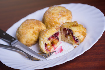 Fresh plum dumplings. One sliced dumpling and two whole with knife and fork on white plate on dark red table