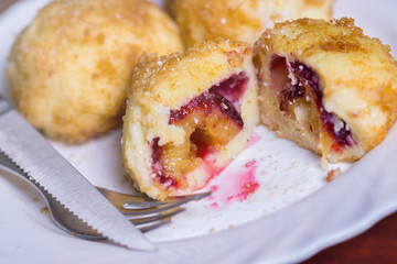 Fresh plum dumplings. One sliced dumpling and two whole with knife and fork on white plate on dark red table
