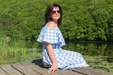 Naklejka premium Young woman in sunglasses sits on the bridge near the river on a sunny day.
