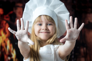 portrait of a little girl in a Baker's hat putting forward his hands in flour on a dark background and a background of fire.