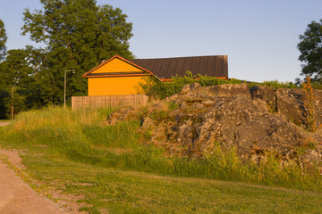 A wooden rural house on the island of Suomenlinna, Sveaborg in Finland and a rural view with a large stone and grass in the summer at sunset.
