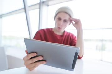 Young man sitting at a table near a window with a tablet in his hands. Young businessman in casual clothing, sitting in a cafe and using a tablet. Tablet is close-up and in focus