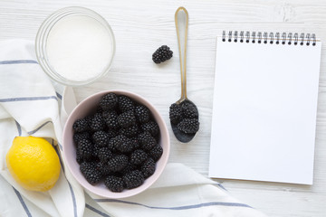 Ingredients for blackberries jam(berries, lemon, sugar) with notepad on a white wooden background. From above, overhead. Space fot text.