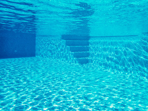 underwater image of an outside villa swimming pool with some steps at one side. The water is clear blue and the sunlight is making patterns