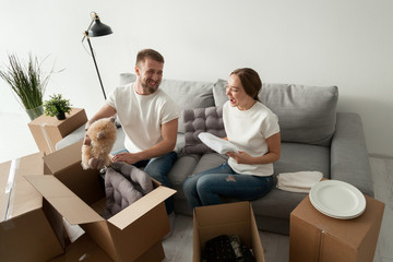Excited husband and wife laughing while unpacking cardboard boxes relocating to new home, young...