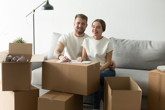 Portrait of happy millennial couple sitting on cozy sofa in new purchased home near cardboard boxes, excited spouses looking at camera glad to move in first shared apartment. Living together concept