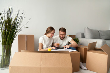 Excited young spouses discussing house blueprint, sitting on floor of newly bought apartment and...