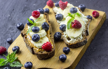 Traditional American and European summer breakfast: sandwiches of toast with peanut butter or cream cheese with raspberries and blueberry, banana. Black concrete table. copy space top view