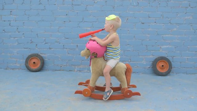 Funny boy blond in superhero mask is sitting on toy rocking horse. child is armed with sword and signal horn and dreams of adventures, travels, exploits. Concept training of spirit, patriotizm