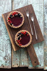 Small vegan tarts made of nuts and berry jam decorated with black, red and white currants on oak cutting board