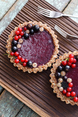 Small vegan tarts made of nuts and berry jam decorated with black, red and white currants on oak cutting board