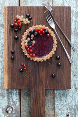 Small vegan tarts made of nuts and berry jam decorated with black, red and white currants on oak cutting board