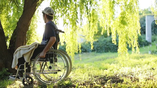 side view of young handsome handicapped man riding on wheelchair in summer park with beautiful sun light shining on background sunny green forest complete physical recovery paralysis rehabilitation