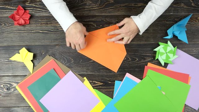 Close up male hands folding origami figure. Top view. Set of colourful papers on wooden table.