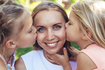 Waist up portrait of smiling woman spending time with children in park. Daughters are kissing her at cheeks while female is laughing in delight